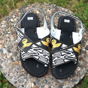 May include: Black leather sandals with white and gold embroidered straps featuring a geometric pattern and a stylized bird design.