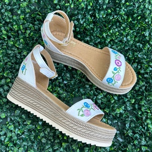 May include: White platform sandals with a floral embroidery design. The sandals have an ankle strap and a woven platform sole.
