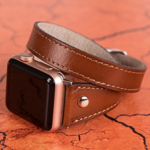 May include: A brown leather watch band with a rose gold Apple Watch. The band is double wrapped and has a stitched edge.
