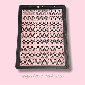 May include: A rectangular black tray containing twenty-four small, light pink rectangular inserts arranged in a grid. Each insert features a repeating cutout pattern of overlapping semicircles. The text "seigaiha | sakura" is printed below.