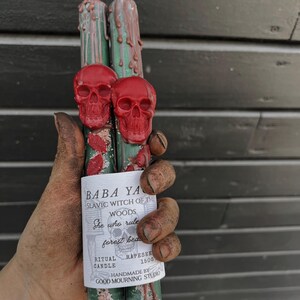 Könnte beinhalten: Zwei dunkelgrüne Stabkerzen mit roten Wachstotenkopfverzierungen und roten Wachstropfen. Ein weißes Papieretikett ist um die Kerzen gewickelt mit dem Text BABA YAGA SLAVIC WITCH OF THE WOODS She who rules forest beast RITUAL CANDLE RAPESEED 1500.