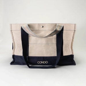 May include: A large, beige canvas tote bag with navy blue trim and straps. The bag has a single pocket on the front and the word "CONDO" is printed in gold on the front.