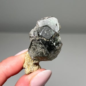 May include: A dark green and black crystal cluster with a rough, natural texture. The crystal is attached to a smaller, light brown rock.
