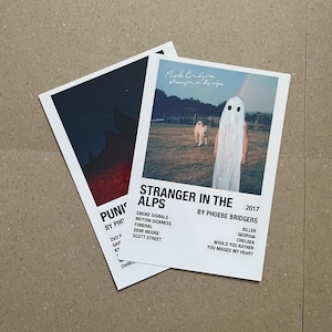 Peut inclure: Une affiche blanche avec du texte noir qui dit "Stranger in the Alps" par Phoebe Bridgers. L'affiche présente une photo d'une personne portant un costume de fantôme blanc et un chien marchant dans un champ.