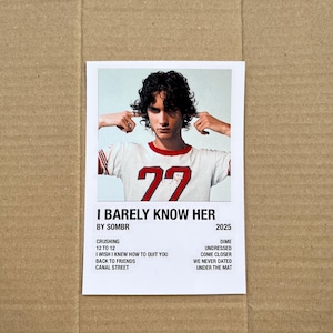 May include: A white and red t-shirt with the number 77 is worn by a person on a white polaroid-style print. The print also includes the text "I BARELY KNOW HER" and a list of song titles. The background is a cardboard surface.