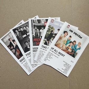 Może przedstawiać: Kolekcja pięciu białych plakatów One Direction. Każdy plakat przedstawia inny obraz członków zespołu, z tytułami piosenek i tekstami wydrukowanymi na czarno. Plakaty są ułożone w kształt wachlarza.