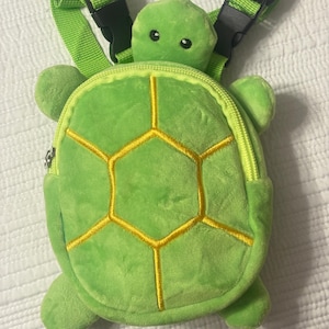 Könnte beinhalten: Ein grüner Rucksack in Schildkrötenform mit einem gelb genähten Schalenmuster. Der Rucksack hat einen Schildkrötenkopf und vier kleine Füße. Er verfügt über verstellbare grüne Träger mit schwarzen Schnallen und ein Reißverschlussfach.
