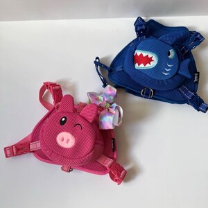 Puede incluir: Dos mochilas infantiles coloridas con correas ajustables. Una mochila presenta una cara de cerdo rosa y la otra una cara de tibur&oacute;n azul.