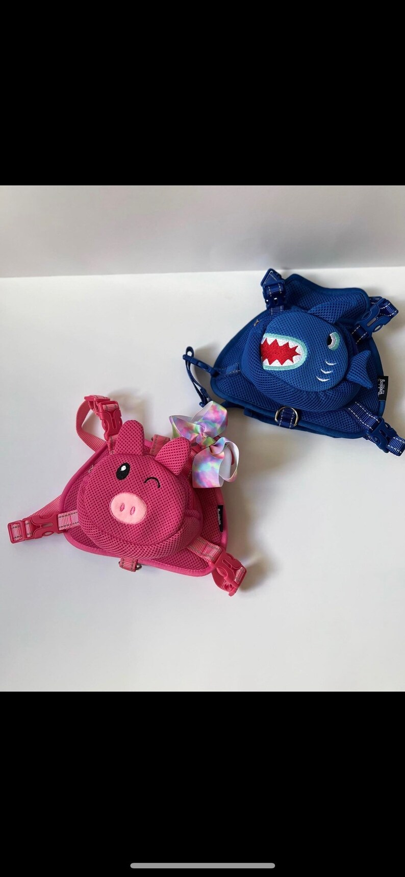 Puede incluir: Dos mochilas infantiles coloridas con correas ajustables. Una mochila presenta una cara de cerdo rosa y la otra una cara de tibur&oacute;n azul.