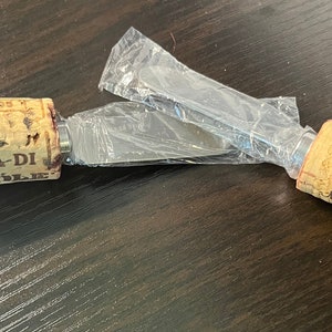 May include: A stainless steel cheese knife with cork handles. The handles are labeled "Tenuta di Mozzale" and "Menage a Trois".