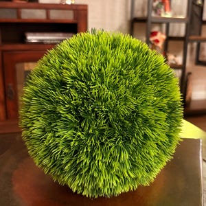 May include: A spherical decorative object with a vibrant green, grass-like texture. The artificial plant is round and sits on a dark brown surface. The object is a decorative accent piece.