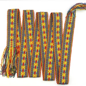 May include: A long, woven textile belt featuring a repeating geometric pattern in vibrant red, yellow, and blue threads. The band is folded in a zigzag formation and finished with loose, multi-colored fringe tassels at both ends of the fabric.