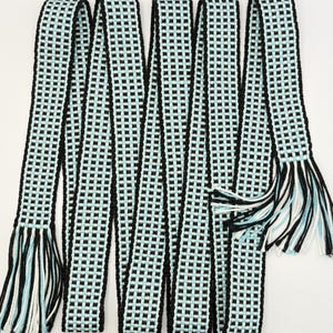 May include: A long, woven belt featuring a repeating checkered pattern in black, white, and light blue. The belt has a flat, narrow construction with solid black edges and ends finished with loose, multi-colored fringe tassels in black, white, and blue.