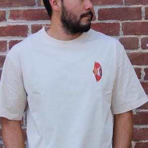 Op de afbeelding: Een beige T-shirt met een rode, oranje en gele afbeelding van een pizzapunt op de voorkant.