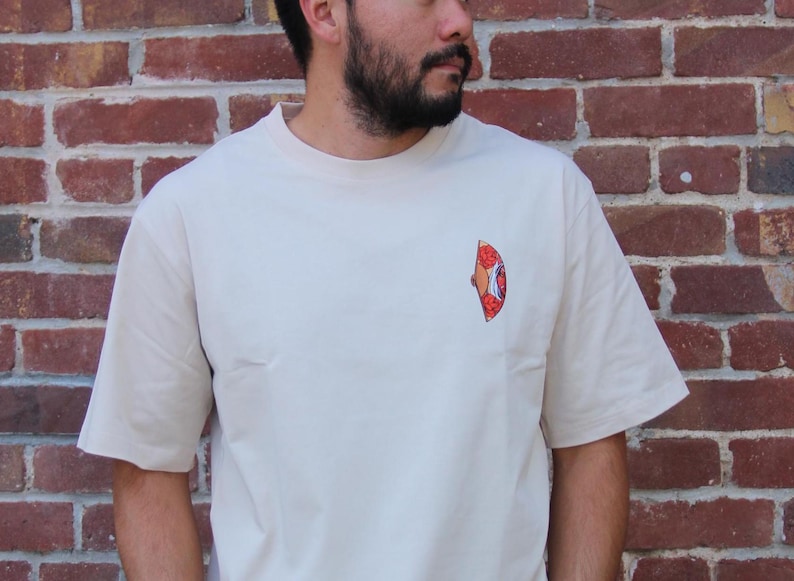 Op de afbeelding: Een beige T-shirt met een rode, oranje en gele afbeelding van een pizzapunt op de voorkant.