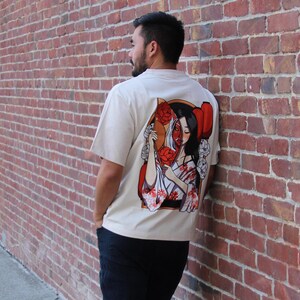 Op de afbeelding: Een wit T-shirt met een grafische print van een vrouw die een waaier vasthoudt. Het gezicht van de vrouw is gedeeltelijk verborgen door de waaier, en ze draagt een traditionele Japanse kimono. De grafiek is in tinten rood, wit en zwart.