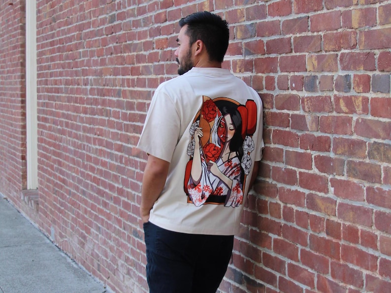 Op de afbeelding: Een wit T-shirt met een grafische print van een vrouw die een waaier vasthoudt. Het gezicht van de vrouw is gedeeltelijk verborgen door de waaier, en ze draagt een traditionele Japanse kimono. De grafiek is in tinten rood, wit en zwart.