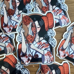 May include: A collection of stickers featuring a geisha-like figure with a split face design, one side human and the other a demon. The geisha wears a white kimono with orange floral patterns and holds a fan. Lanterns and flowers surround the central figure.