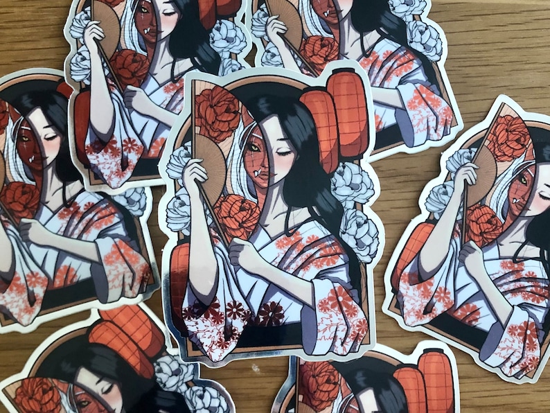 May include: A collection of stickers featuring a geisha-like figure with a split face design, one side human and the other a demon. The geisha wears a white kimono with orange floral patterns and holds a fan. Lanterns and flowers surround the central figure.