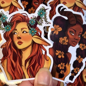 May include: Two illustrated stickers of women with animal features. One woman has red hair and deer ears with blue flowers in her hair. The other woman has dark brown hair and horns with yellow flowers in her hair.