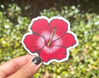 May include: A red hibiscus flower sticker with a white center and yellow stamen. The sticker is on a white background and is being held by a hand.