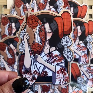 May include: A sticker featuring a geisha-style illustration. The design showcases a woman in a kimono, holding a fan, with a split face revealing a demon mask. Red lanterns and floral elements complete the Japanese-inspired artwork. The sticker has a gold border.