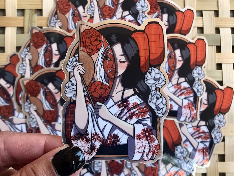 May include: A sticker featuring a geisha-style illustration. The design showcases a woman in a kimono, holding a fan, with a split face revealing a demon mask. Red lanterns and floral elements complete the Japanese-inspired artwork. The sticker has a gold border.