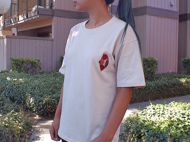 Op de afbeelding: Een wit T-shirt met een rood en zwart grafisch ontwerp aan de linkerkant van de borst. Het grafische ontwerp toont een gestileerde afbeelding van een waaier.
