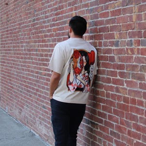 Op de afbeelding: Een wit T-shirt met een grafische print van een vrouw met rode bloemen in haar haar. De vrouw draagt een witte kimono met rode bloemen. De grafiek bevindt zich op de achterkant van het shirt.