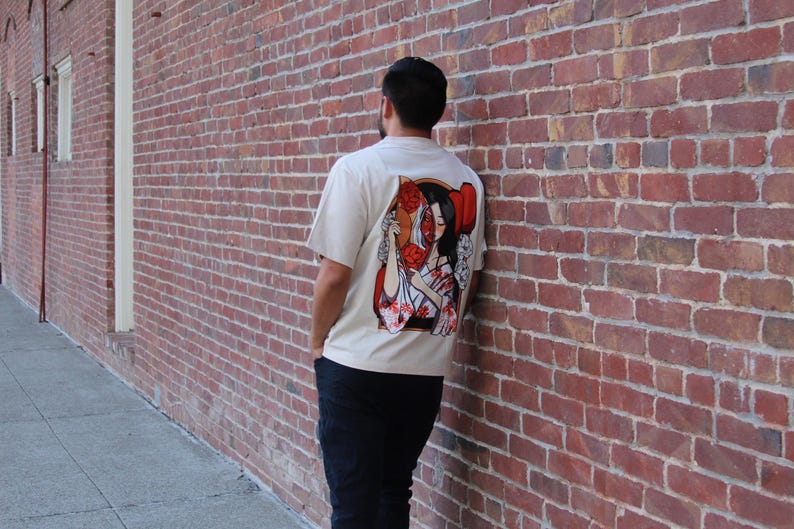 Op de afbeelding: Een wit T-shirt met een grafische print van een vrouw met rode bloemen in haar haar. De vrouw draagt een witte kimono met rode bloemen. De grafiek bevindt zich op de achterkant van het shirt.