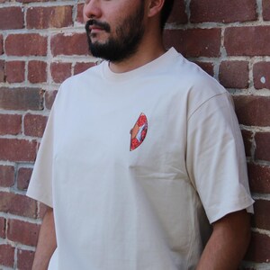 Op de afbeelding: Een beige T-shirt met een kleine, kleurrijke afbeelding op de voorkant. De afbeelding is een gestileerde afbeelding van een vis.