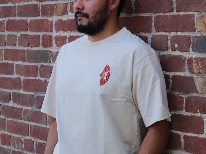 Op de afbeelding: Een beige T-shirt met een kleine, kleurrijke afbeelding op de voorkant. De afbeelding is een gestileerde afbeelding van een vis.
