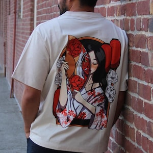Op de afbeelding: Een wit T-shirt met een grafische print van een vrouw die een rode en witte waaier vasthoudt, met bloemen en een rood masker dat haar gezicht gedeeltelijk bedekt.
