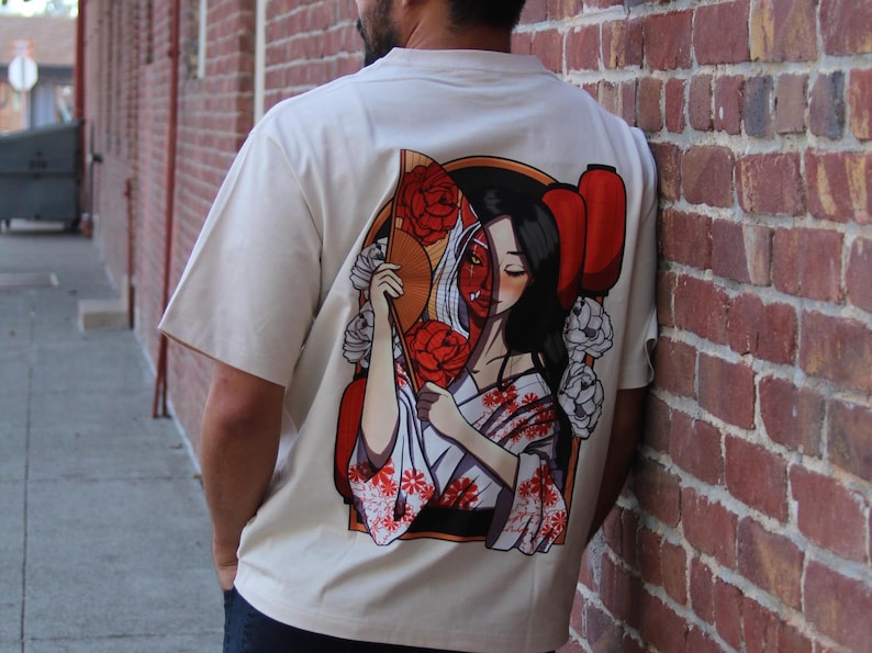 Op de afbeelding: Een wit T-shirt met een grafische print van een vrouw die een rode en witte waaier vasthoudt, met bloemen en een rood masker dat haar gezicht gedeeltelijk bedekt.