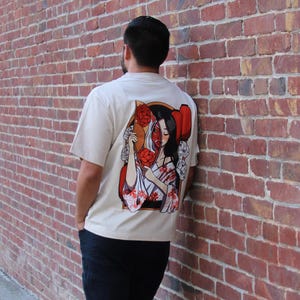 Op de afbeelding: Een wit T-shirt met een grafische print van een vrouw met rode bloemen en een waaier. Het gezicht van de vrouw is gedeeltelijk bedekt door de waaier.