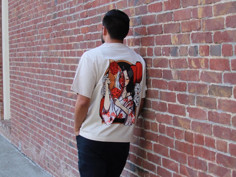 Op de afbeelding: Een wit T-shirt met een grafische print van een vrouw met rode bloemen en een waaier. Het gezicht van de vrouw is gedeeltelijk bedekt door de waaier.