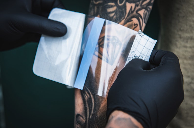 May include: A person's arm with a tattoo is being covered with a clear, protective film. The film is being applied by a gloved hand.