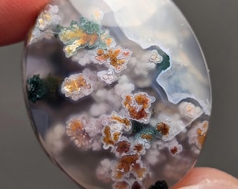 May include: A polished agate cabochon with a white, gray, and brown background. The stone has a unique pattern of green, brown, and white dendrites.