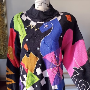 May include: A black turtleneck sweater with a vibrant, abstract design. The sweater features patches of red, green, blue, pink, and orange, with embroidered details and sparkling embellishments. The design includes geometric shapes and swirling patterns.