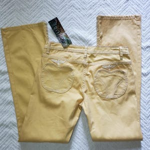 May include: Mustard yellow pants featuring a straight-leg cut and a high-rise waist. The back pockets are shaped like apples with white embroidered outlines and stems. A rectangular brand tag labeled REVOLT is attached to the waistband.