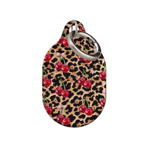 Könnte beinhalten: Ein tropfenförmiger Schlüsselanhänger mit Leopardenmuster und roten Kirschillustrationen. Der Schlüsselanhänger hat einen silberfarbenen Metallring zur Befestigung. Das Design zeigt ein sich wiederholendes Muster aus Kirschen und Leopardenflecken.