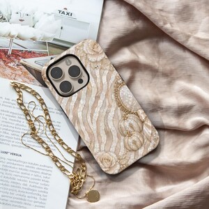 May include: A beige and cream phone case featuring a textured zebra stripe pattern, embellished with decorative gold-toned pumpkins, floral accents, and a dangling gold-toned chain strap attached to the side of the protective cover.
