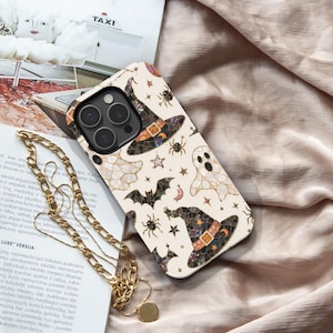 May include: A smartphone case with a cream background featuring a mosaic-style pattern of black witch hats, white ghosts, black bats, spiders, and small stars. The case has a black camera cutout area and is accompanied by a gold-toned chain necklace.