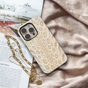 May include: A smartphone case featuring a light beige and white leopard print pattern with a glossy finish. The case has a square camera cutout with three circular lenses. A gold-toned metal chain necklace with flat links rests on top of the case.