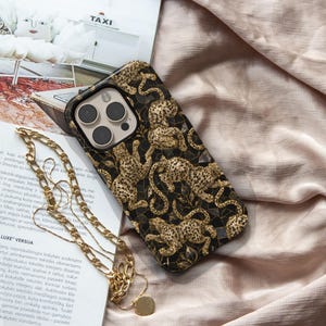 May include: A black smartphone case featuring a repeating pattern of gold-coloured leopards amidst dark foliage and geometric shapes. The case has a cutout for a triple-lens camera system and is accompanied by a gold-toned metal chain strap.