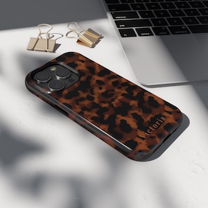 Może przedstawiać: Brązowo-czarne etui na telefon z wzorem w szylkret, z nadrukiem "Victoria". Etui ma błyszczące wykończenie. Dwa złote spinacze i laptop w tle.