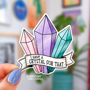 A sticker in the shape of three pastel shaded crystals in pink, purple, and turquoise. At the bottom is a white banner with black text that reads "I have a crystal for that"