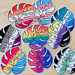 May include: Nine colourful monstera leaf stickers with different pride flag designs. The stickers are printed on white vinyl and have a glossy finish.