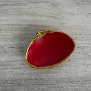 May include: A small red and gold dish made of clay. The dish is shaped like a seashell and has a gold rim.