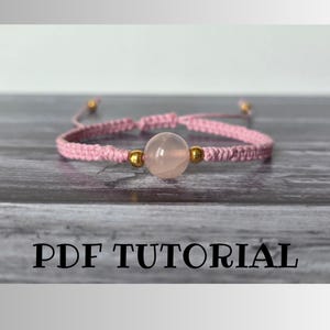 Può includere: Un braccialetto intrecciato rosa con un unico ciondolo rotondo di pietra rosa chiaro e dettagli dorati. Il braccialetto è su una superficie di legno. PDF TUTORIAL è scritto in testo nero sotto il braccialetto.